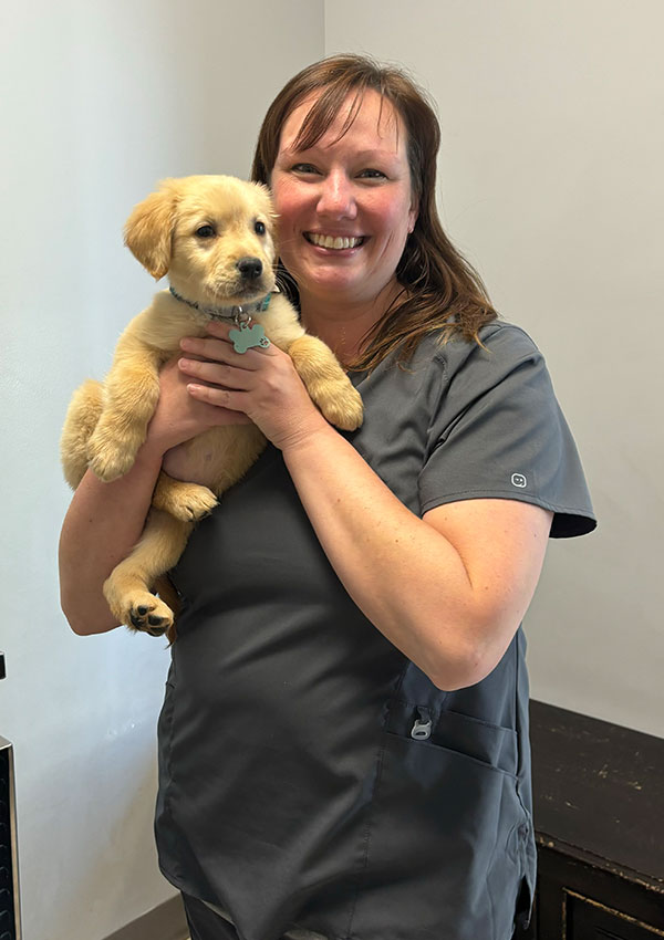 A smiling woman in gray scrubs holds a small, light-colored puppy indoors, standing against a plain light-colored wall.