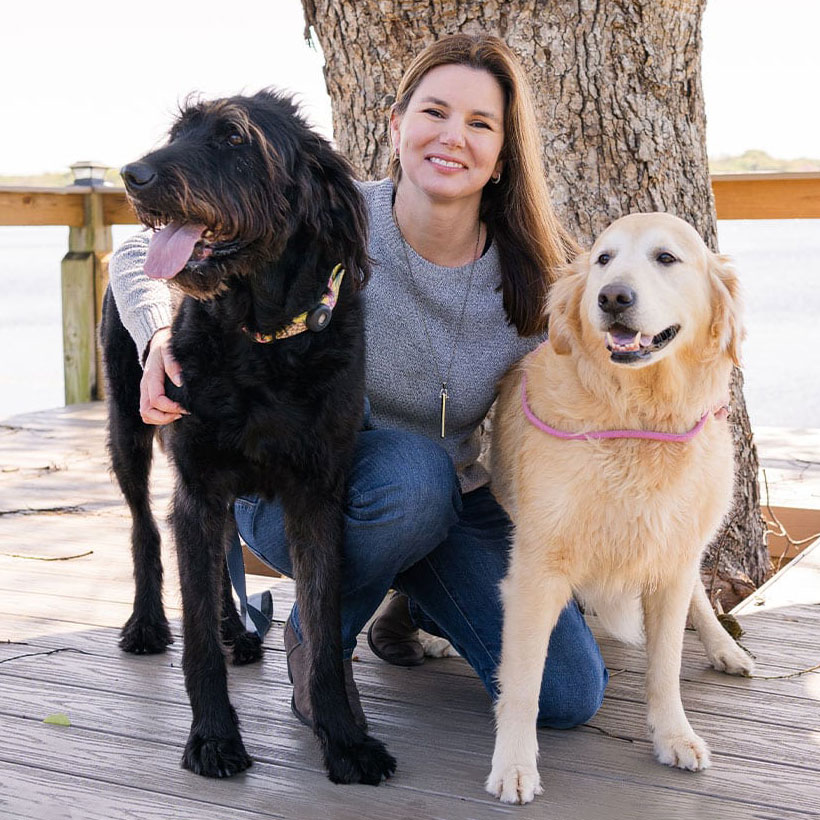 A woman kneels outdoors by a large tree, smiling and posing with a fluffy black dog and a golden dog on a wooden deck near the water.